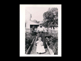 Woman and child (Minnie Taylor and Dizzy) along path in garden with dog. Part of a frame house ("Holmefield") is in the background.